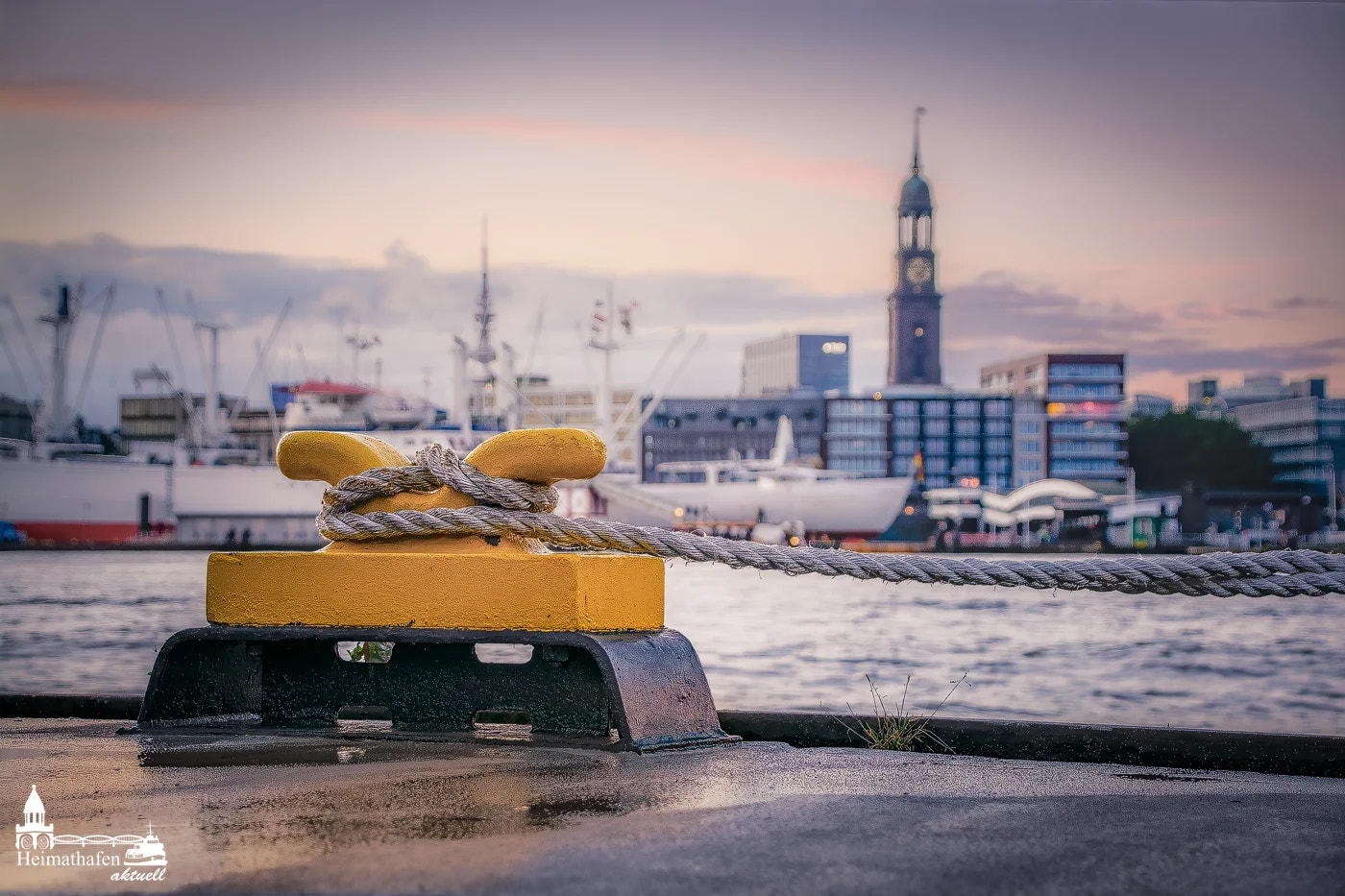 Ein gelber Poller mit festgebundenem Tauwerk im Vordergrund, im Hintergrund die Cap San Diego und der Hamburger Michel bei Abendlicht.