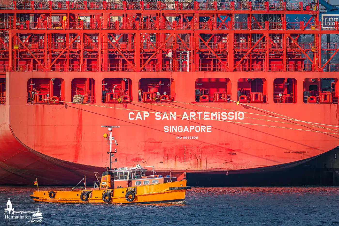 Gelbes Wasserboot fährt vor dem knallroten Riesen-Containerschiff CAP SAN ARTEMISSIO im Hamburger Hafen vorbei.