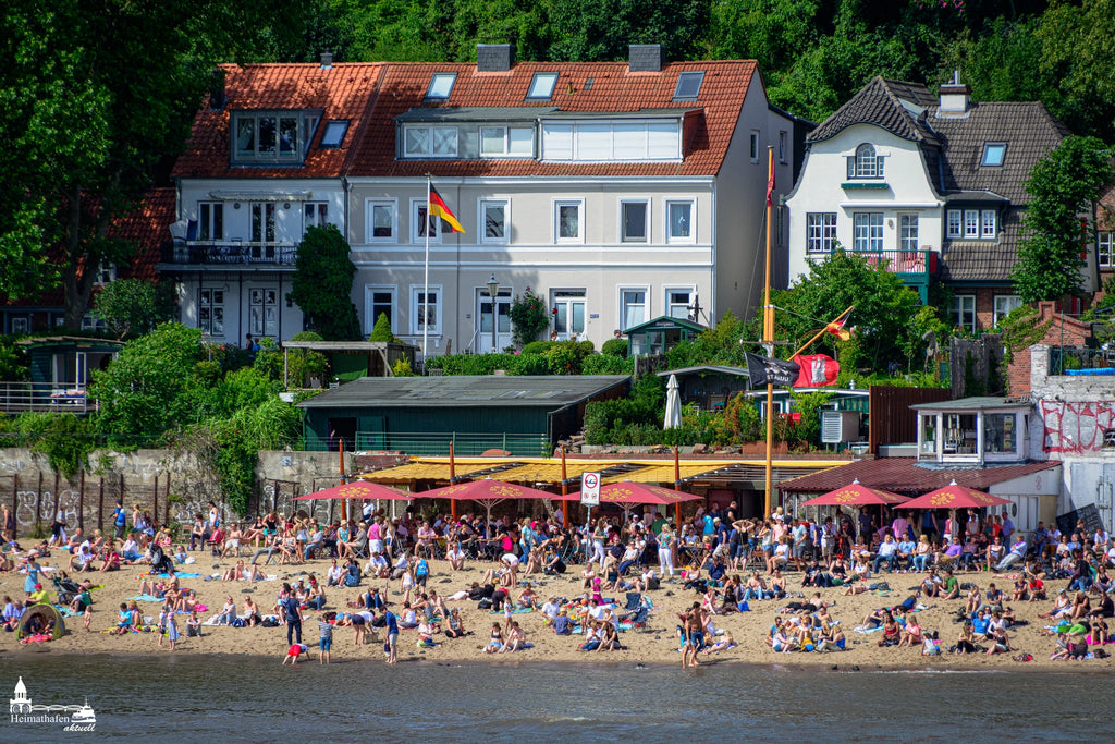 Elbstrand Hamburg im Sommer mit vielen Menschen am Sandstrand und historischen Häusern im Hintergrund.