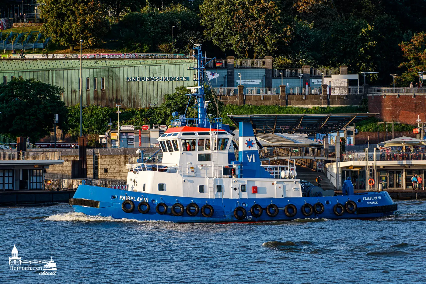 Der Schlepper FAIRPLAY VI aus Rostock fährt vor den Landungsbrücken in Hamburg entlang. Im Hintergrund sind die bekannten grünen Kupferdächer der Station zu sehen.
