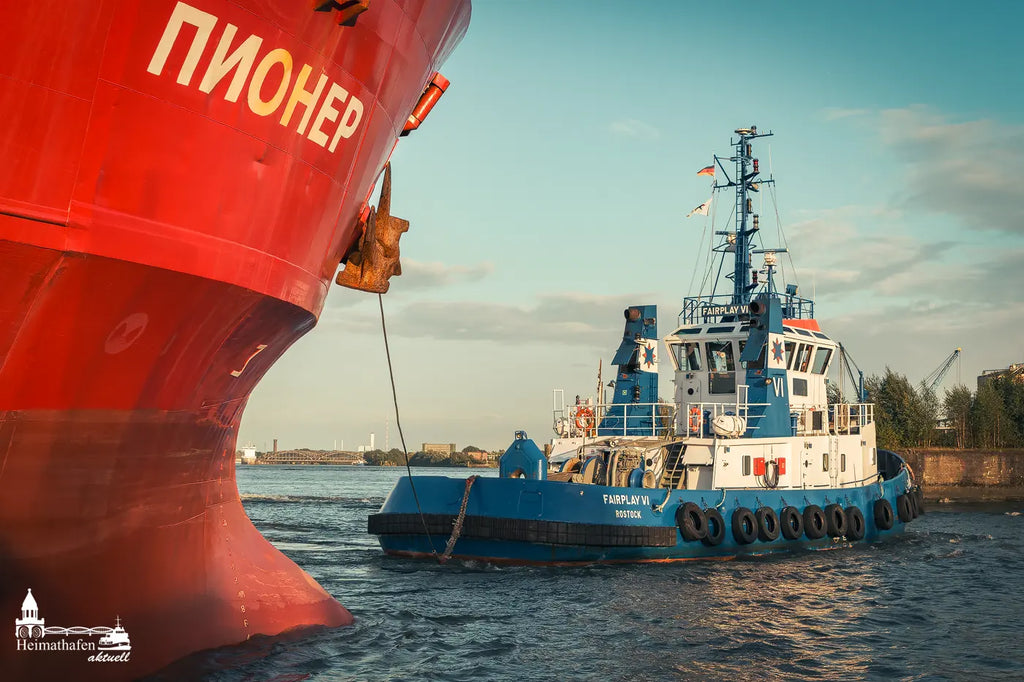 Der blaue Hafenschlepper FAIRPLAY VI aus Rostock zieht ein riesiges Frachtschiff mit kyrillischer Beschriftung ("ПИОНЕР") im Hamburger Hafen bei klarem Abendlicht.